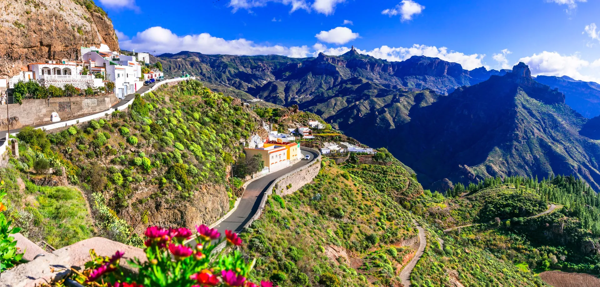 Houses and a winding road on the cliff at Artenara, with Roque Bentayga and Roque Nublo across the caldera