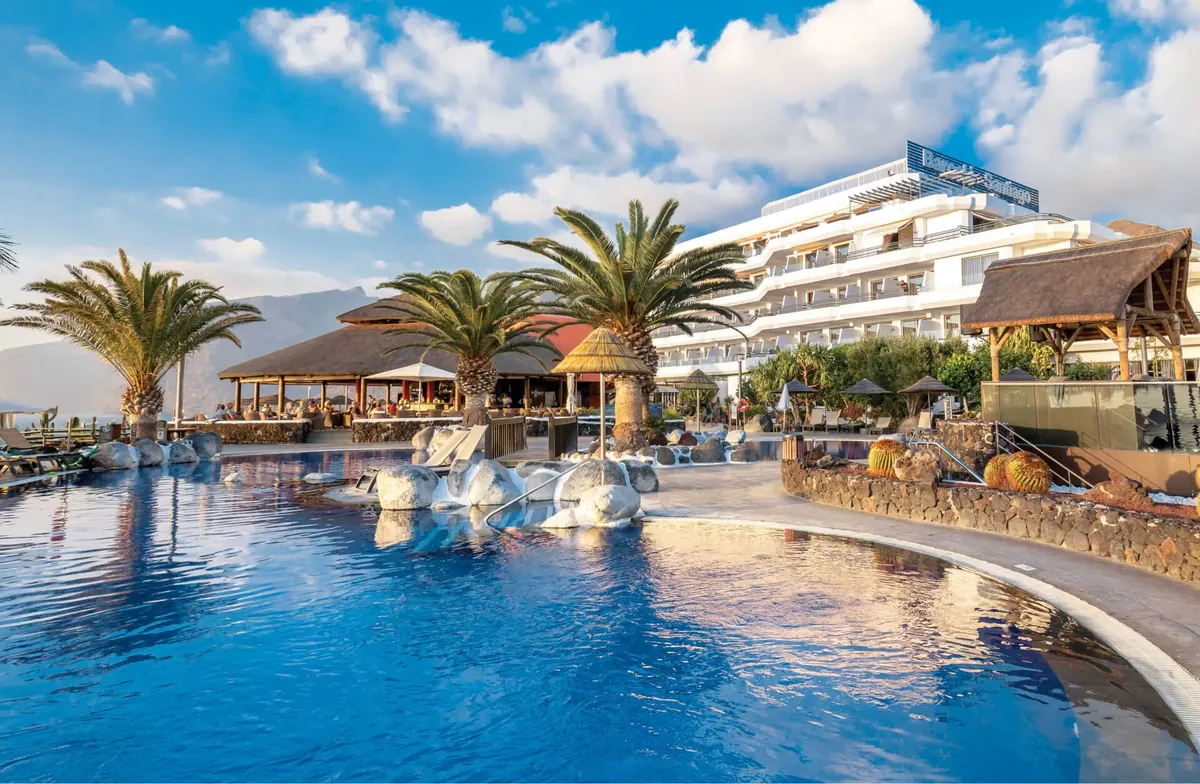 Pool, palm trees and cliff-top building of Barceló Santiago adults-only hotel with Los Gigantes behind