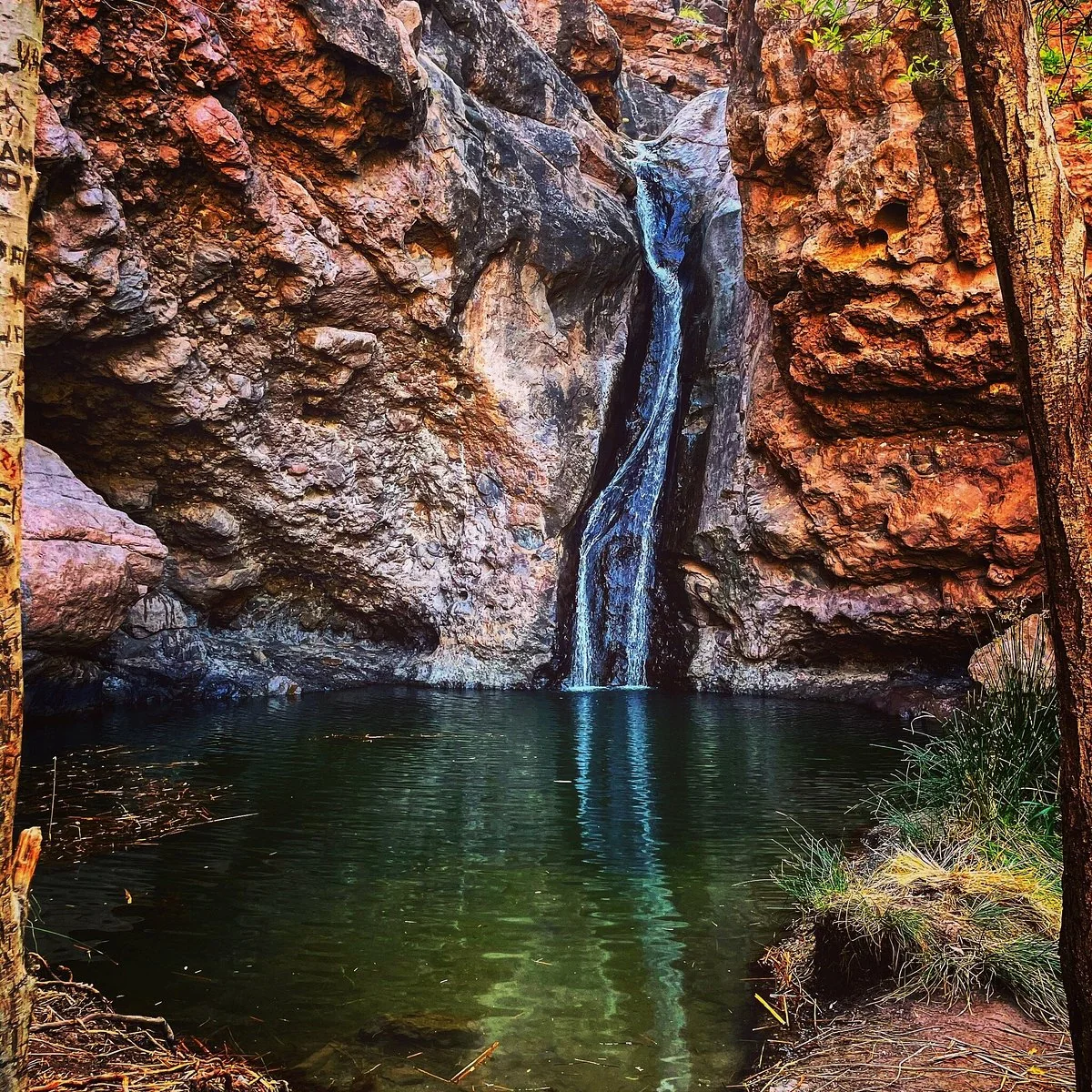 Waterfall falling into the green pool of the Charco Azul, framed by red rock walls in El Risco de Agaete